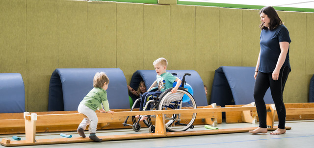 Ein Kind im Rollstuhl und ein Kleinkind spielen gemeinsam auf einer Turnbank in einer Sporthalle
