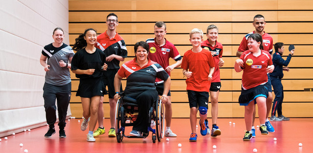 Gruppe von Kindern und Jugendlichen, darunter ein Rollstuhlfahrer, beim gemeinsamen Sport in einer Halle mit Tischtennisbällen auf dem Boden.